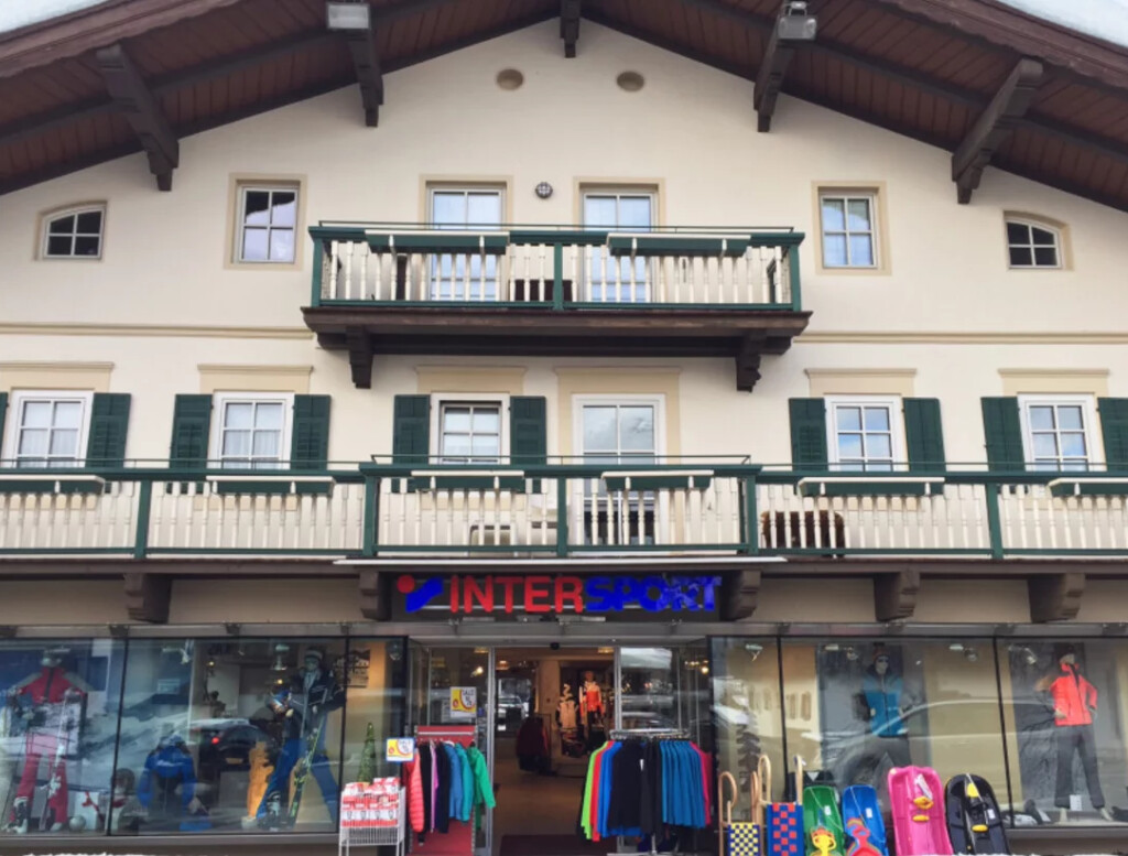 Fassade eines zweistöckigen Gebäudes mit einem Holzbalkon, an dem das Intersport-Ladenschild angebracht ist. In den Schaufenstern sind farbenfrohe Sportbekleidung ausgestellt.