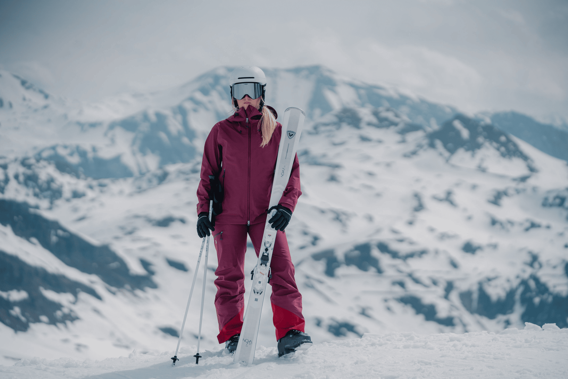 Ein Skifahrer in orangefarbener Kleidung und roter Skibrille steht auf einem schneebedeckten Berg und hält seine Skier über die Schulter. Daneben sind vier Paar Skier senkrecht aufgestellt.