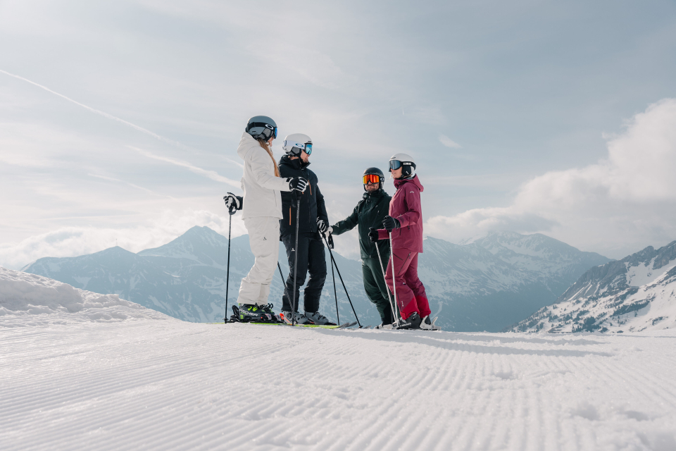 Vier Skifahrer in farbenfroher Ausrüstung stehen mit Skistöcken auf einem verschneiten Berghang. Majestätische Gipfel und ein bewölkter Himmel bilden die ruhige Kulisse.
