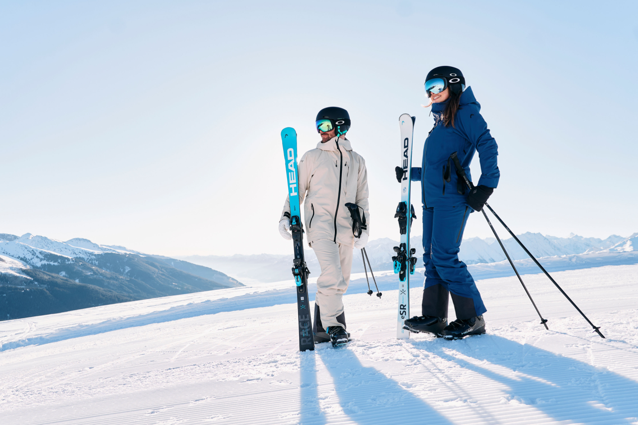 Two skiers wearing helmets and ski gear are standing on a sunlit snowy slope, holding their skis in their hands. In the background, a clear blue sky and distant mountains can be seen.