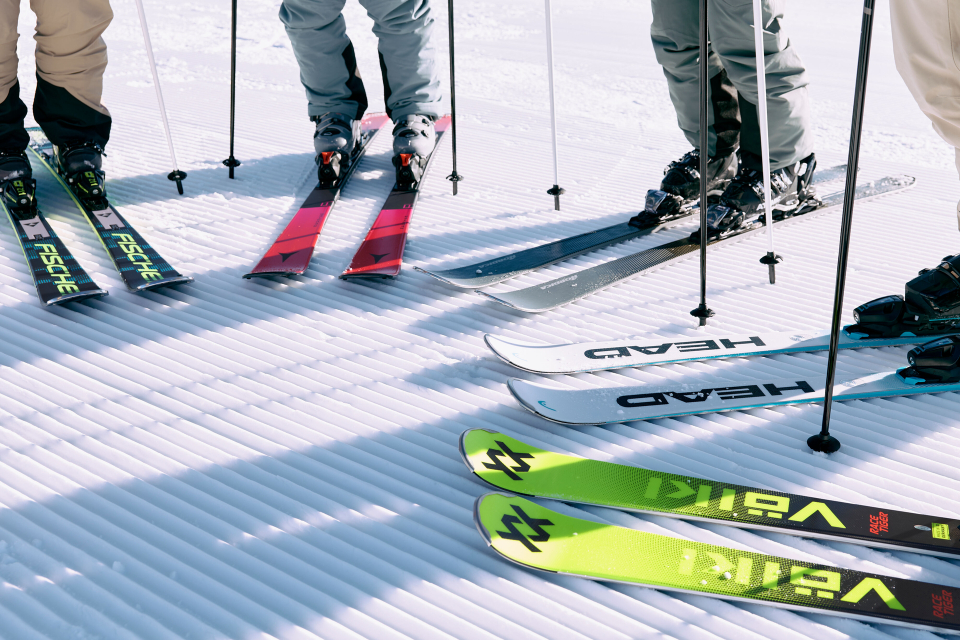 Six pairs of skis with ski poles stand on a sunlit, grooved snow surface. The skis are brightly colored, featuring brands like Völkl and Head.