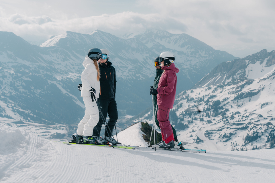 Three skiers in colorful gear stand on a snow-covered mountainside, with majestic peaks towering in the background. The scene conveys camaraderie and a spirit of adventure.