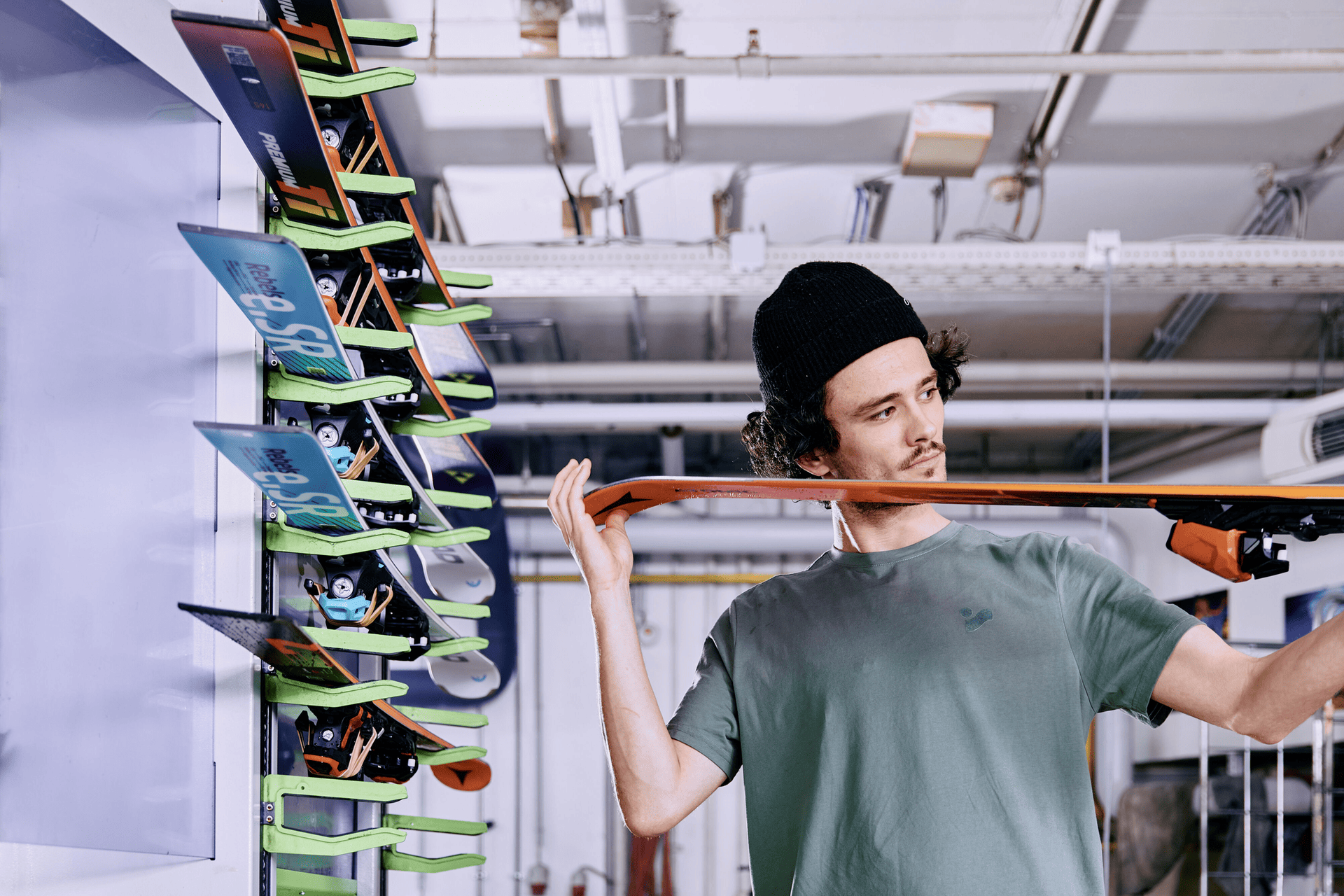 Eine Person mit einer Mütze untersucht ein Snowboard in einer mit Werkzeugen gefüllten Werkstatt genau. Im Hintergrund sind mehrere bunte Snowboards ausgestellt.