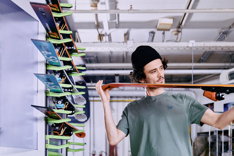 A person wearing a cap carefully examines a snowboard in a workshop filled with tools. Several colorful snowboards are displayed in the background.