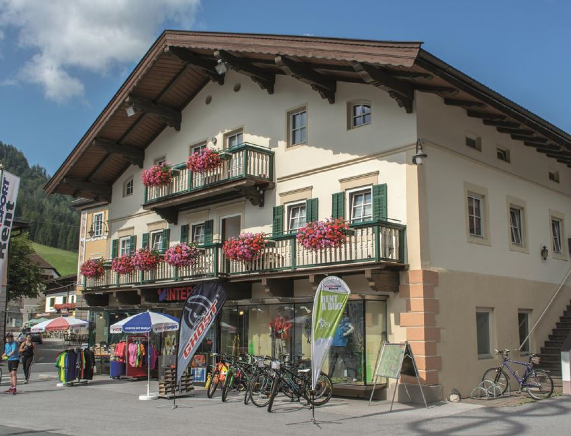 Gebäude im alpinen Stil mit Balkonen, Blumenkästen und Schaufenstern, in denen Sportausrüstung ausgestellt ist. Vor dem Gebäude stehen Fahrräder. Die Szene ist lebhaft und sonnig.