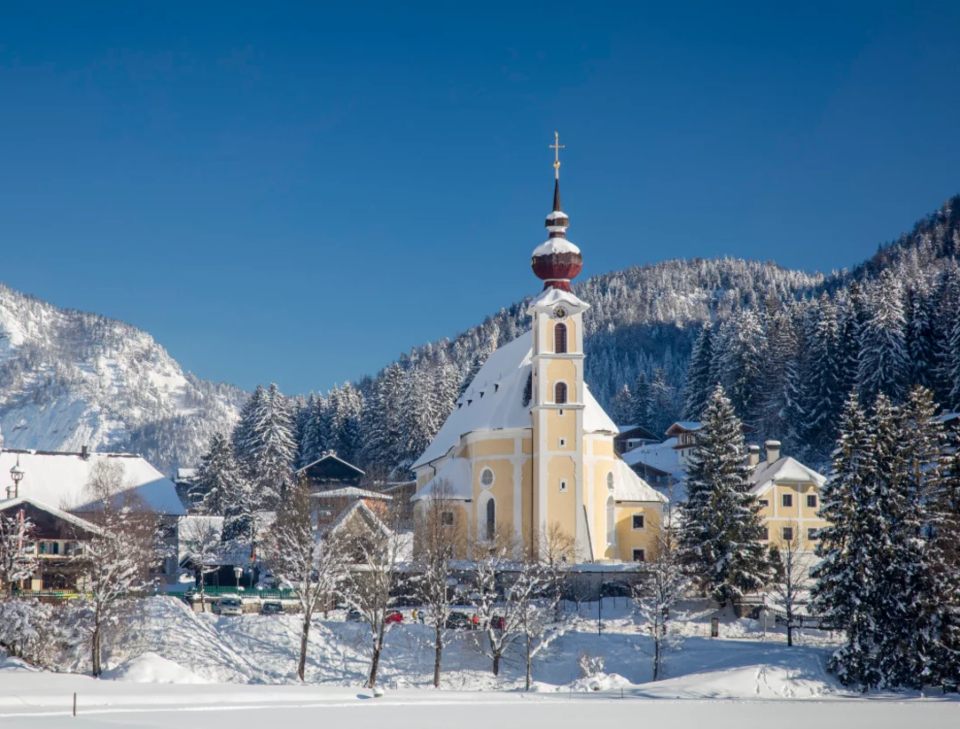 Eine gelbe Kirche mit einer roten Kuppel steht inmitten schneebedeckter Bäume und Berge unter einem klaren blauen Himmel und vermittelt ein ruhiges Winterbild.