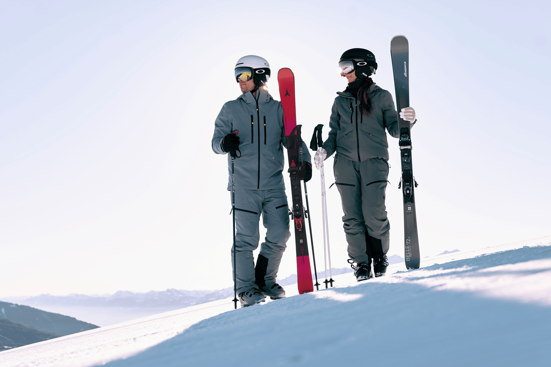 Zwei Skifahrer in grauer Kleidung laufen unter einem strahlend blauen Himmel mit Skiern und Stöcken auf einem schneebedeckten Berg bergauf. Sie wirken entspannt und bereit.