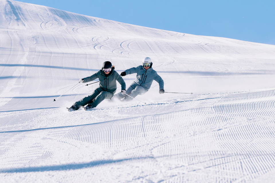 Two skiers in winter gear carve through fresh, groomed snow on a sunny day, casting long shadows. The scene conveys excitement and speed.