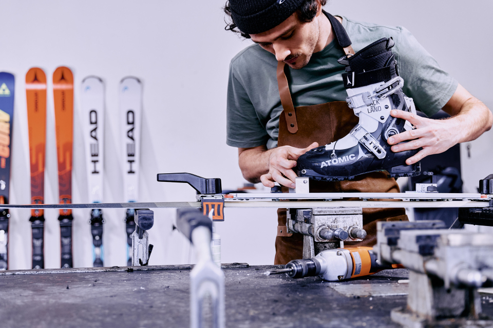 A technician wearing an apron adjusts a ski boot on a workbench surrounded by tools. Skis are lined up in the background, suggesting a workshop environment.