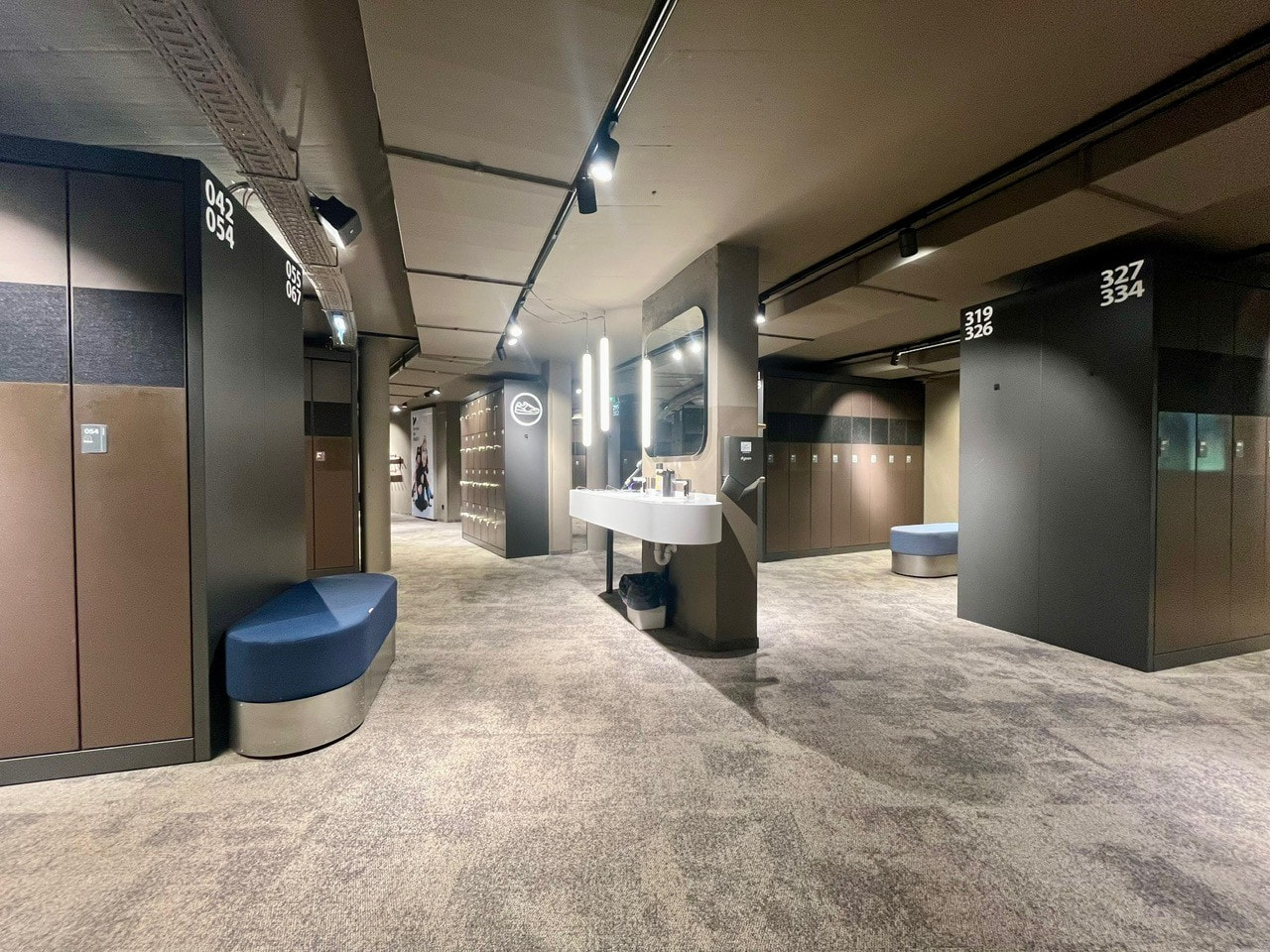 Modern locker room interior with gray carpet, lockers, sink, and bench seating.
