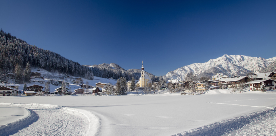 Verschneiter Ort mit Kirche, umgeben von Bergen und winterlichen Wäldern.