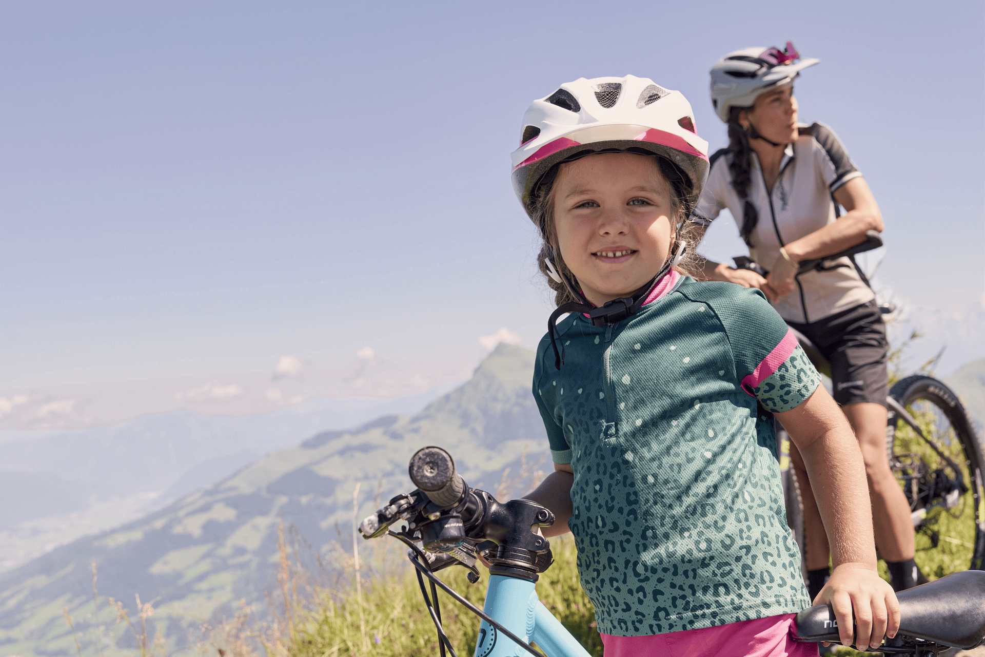 A smiling child wearing a helmet holds a bicycle on a picturesque mountain path. An adult cyclist stands next to them, both enjoying a sunny day with a sweeping view of the valley.