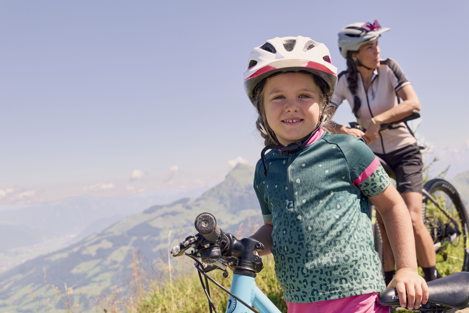 A smiling child wearing a helmet holds a bicycle on a picturesque mountain path. An adult cyclist stands next to them, both enjoying a sunny day with a sweeping view of the valley.