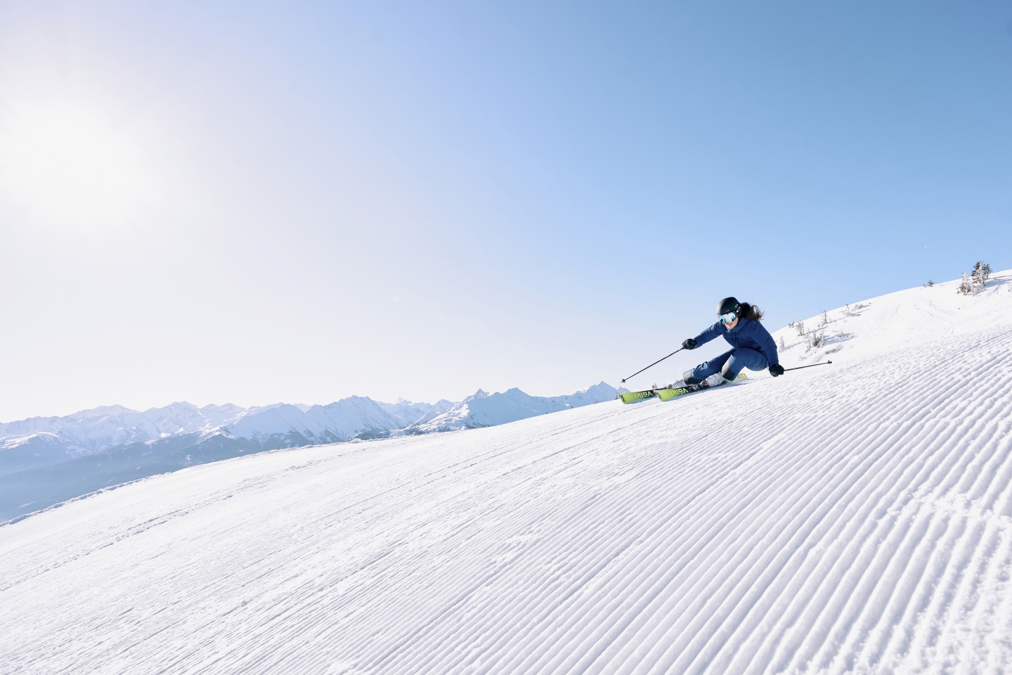 Skifahrer in blauem Anzug fährt auf präparierter Piste mit Bergkette im Hintergrund unter klarem Himmel bergab.