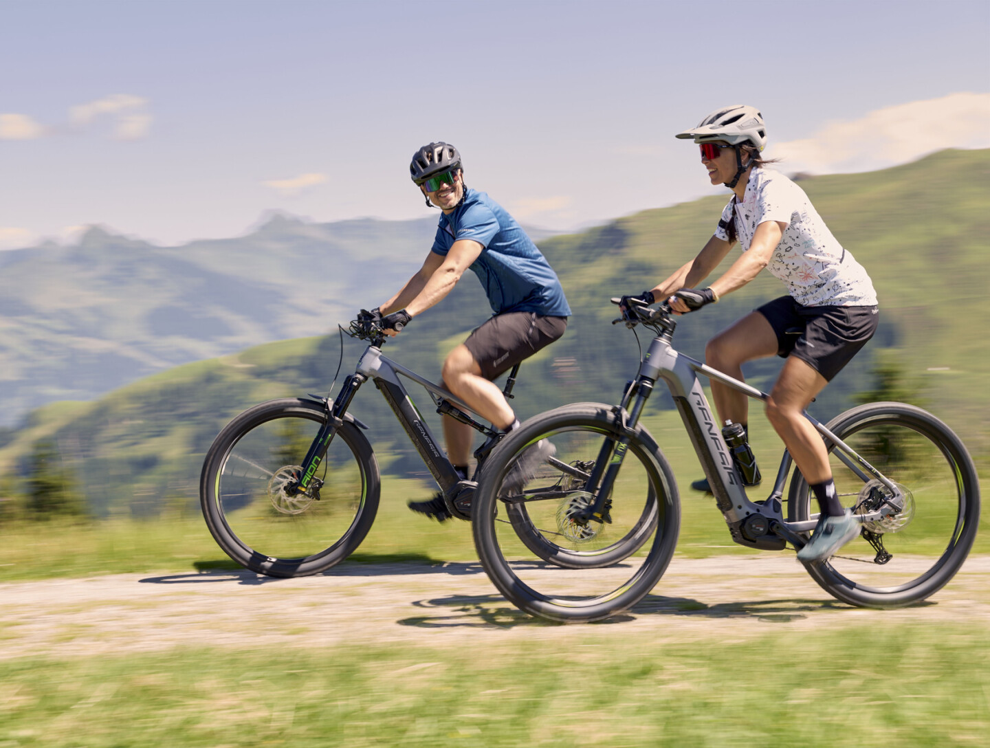Two cyclists on electric mountain bikes riding a sunny mountain trail with blurred peaks.