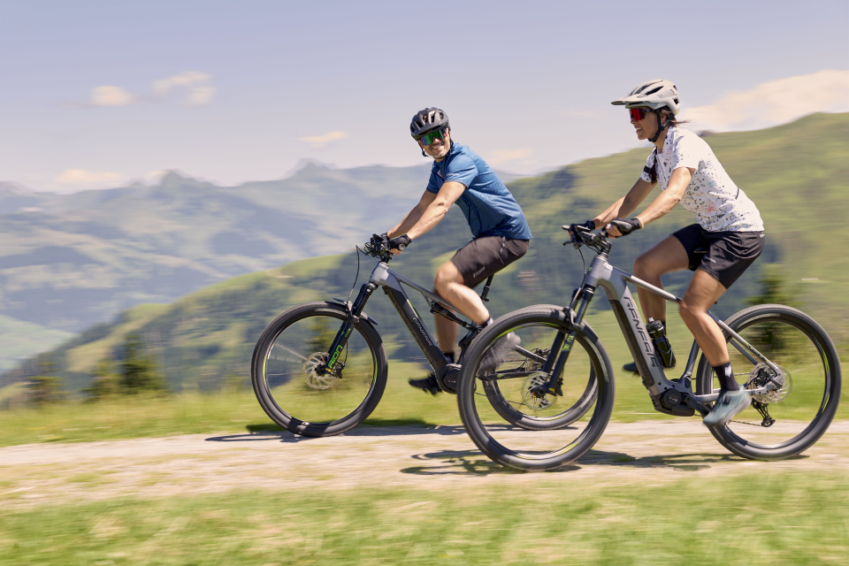 Zwei Radfahrer fahren mit ihren Elektro-Mountainbikes auf einem malerischen Weg durch sanfte grüne Hügel und vorbei an fernen Bergen unter einem klaren, sonnigen Himmel und vermitteln dabei ein Gefühl von Abenteuer und Freude.