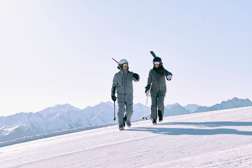 Two skiers in gray outfits and helmets are walking up a snowy slope under a clear blue sky, carrying their skis. Snow-covered mountains can be seen in the background.
