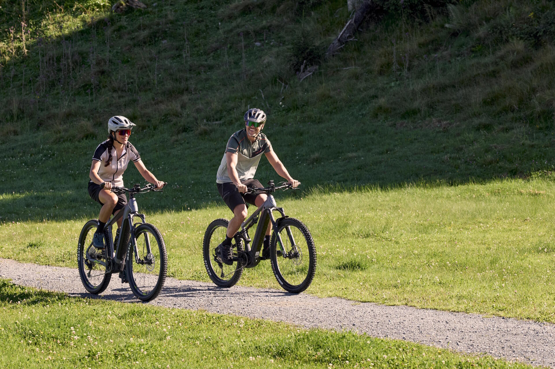Zwei Radfahrer mit Helmen fahren auf E-Mountainbikes einen sonnigen Schotterweg durch grünes Gras.