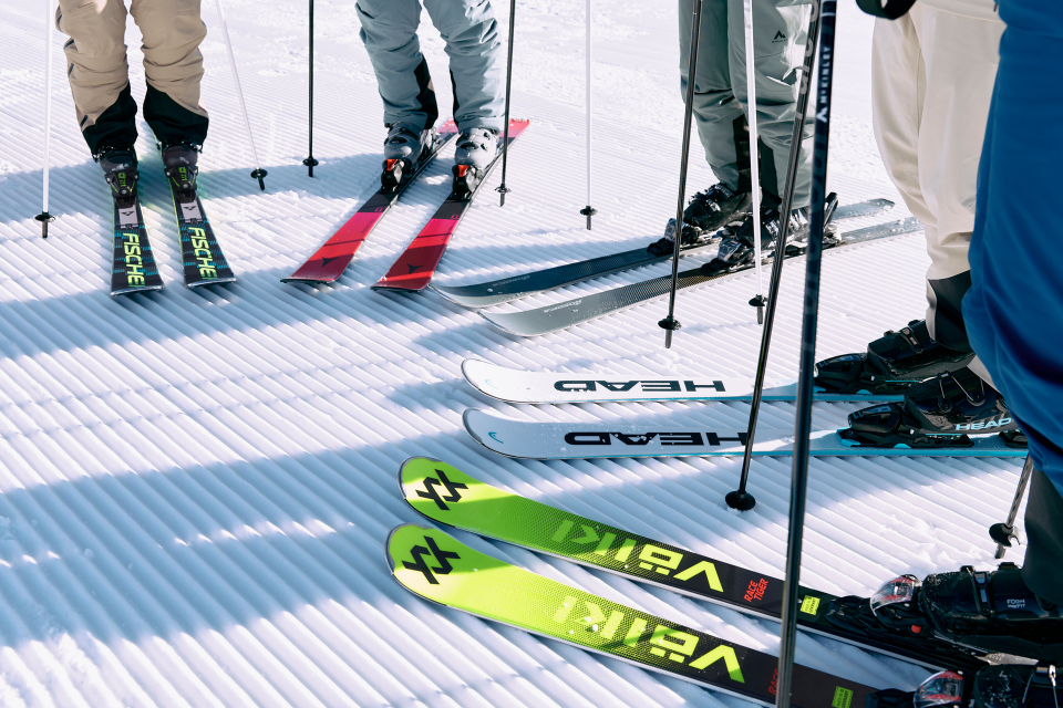 A group of skiers stands on a sunlit, furrowed snow slope, their colorful skis and poles arranged in a circle, ready for the descent.