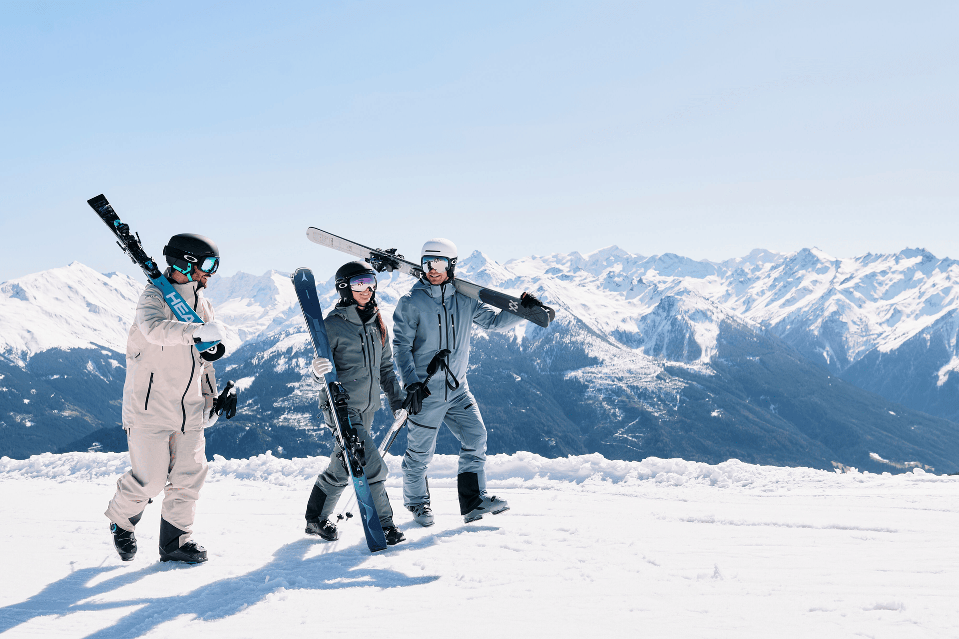 Three people in ski gear are walking on a snowy slope carrying snowboards, while snow-covered mountains stand out against a bright blue sky in the background.