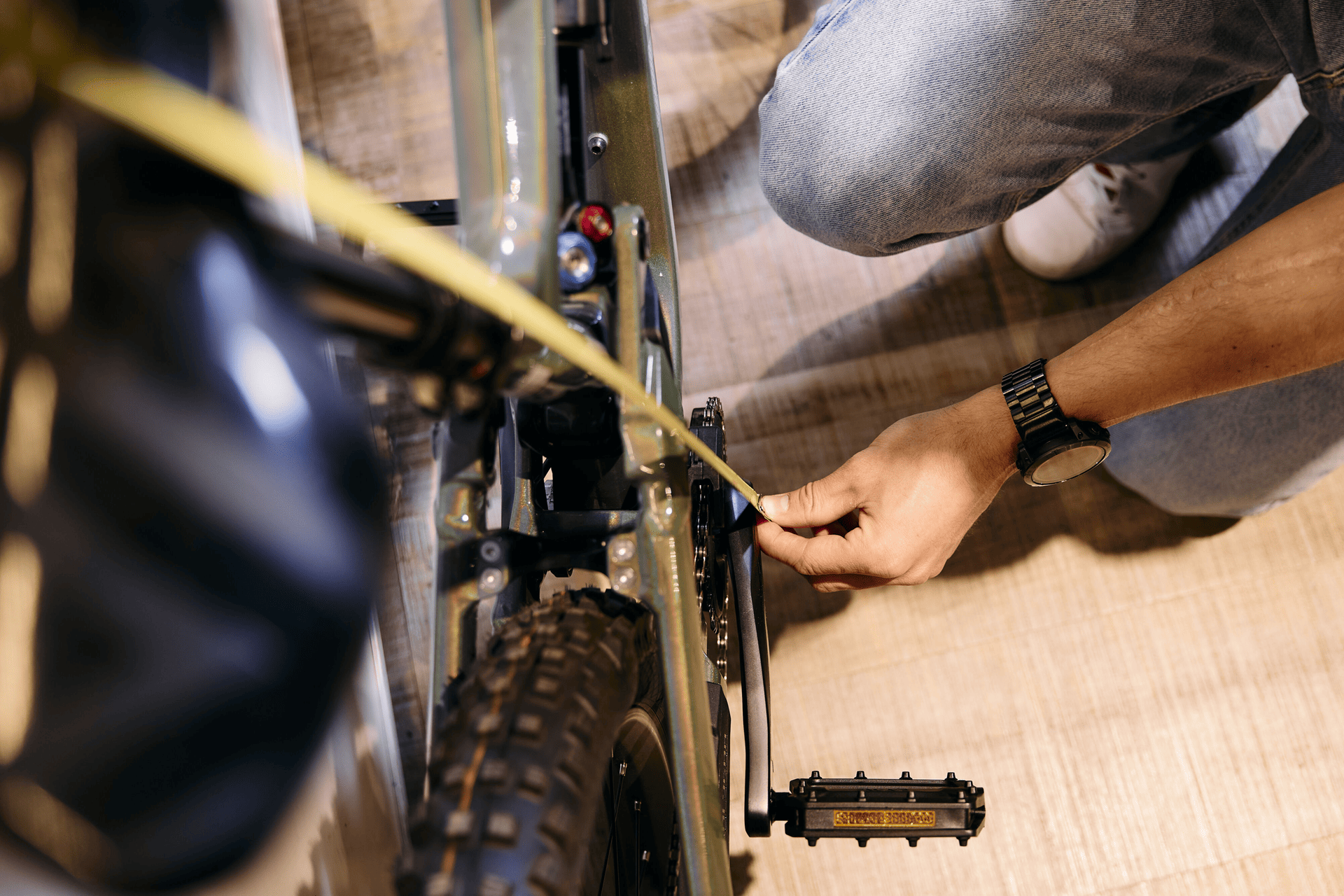 Eine Person in Jeans misst mit einem Maßband auf einem Holzboden den Rahmen eines Fahrrads und achtet dabei auf Präzision. Die Szene ist detailreich und konzentriert.