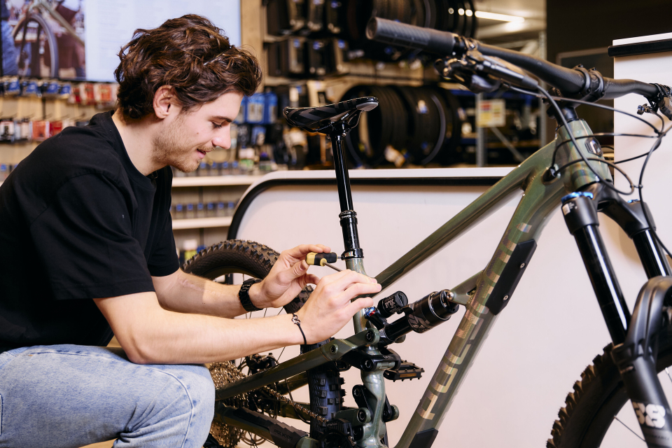 Ein Mann in schwarzem Hemd und Jeans stimmt in einem Fahrradgeschäft konzentriert ein grünes Mountainbike. Im Hintergrund sind Regale mit Reifen und Werkzeugen verschwommen zu sehen.