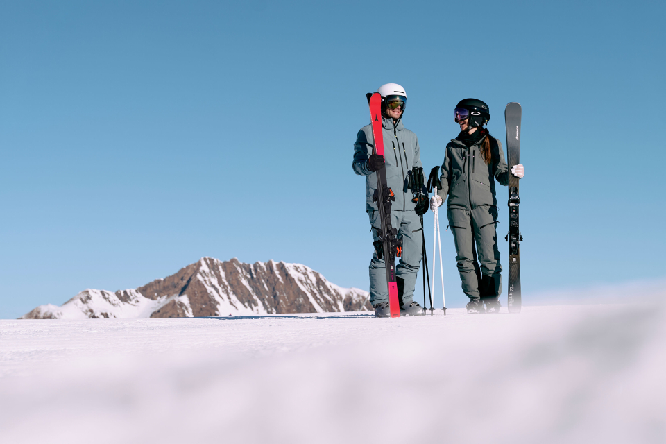 Zwei Skifahrer in grauen Outfits stehen auf einem verschneiten Hang unter einem strahlend blauen Himmel und halten ihre Skier in den Händen. Im Hintergrund sind schneebedeckte Berggipfel zu sehen.