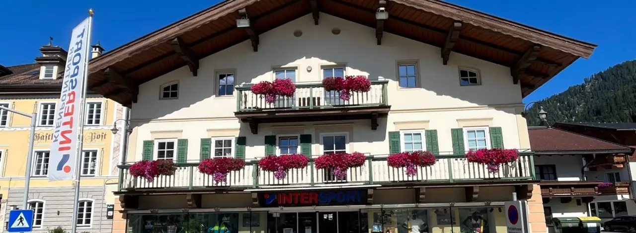 Gebäude im alpinen Stil mit grünen Fensterläden, in dem sich ein Intersport-Geschäft befindet. Die Schaufensterauslage zeigt farbenfrohe Sportbekleidung und -ausrüstung und schafft eine einladende Atmosphäre.