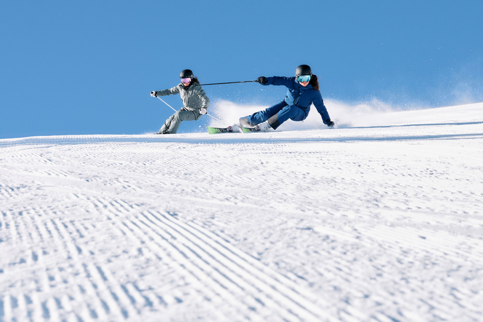 Zwei Skifahrer in Winterausrüstung fahren schnell einen schneebedeckten Hang hinunter vor einem klaren blauen Himmel. Sie beugen sich nach vorne und erzeugen so eine dynamische Bewegung und Spannung.