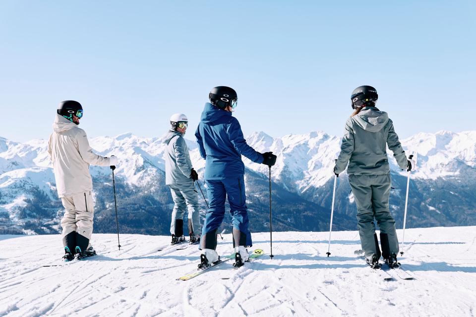 Vier Personen in Skibekleidung stehen mit Skistöcken auf einem schneebedeckten Berg und blicken unter einem klaren blauen Himmel auf die fernen schneebedeckten Gipfel, was ein Gefühl von Abenteuer und Gelassenheit vermittelt.