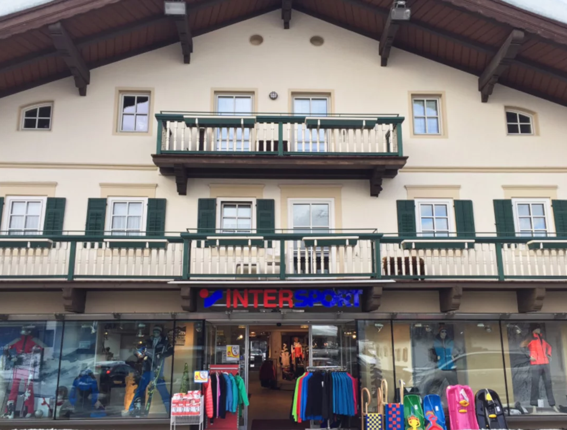 Gebäude im alpinen Stil mit grünen Fensterläden, in dem sich ein Intersport-Geschäft befindet. Die Schaufensterauslage zeigt farbenfrohe Sportbekleidung und -ausrüstung und schafft eine einladende Atmosphäre.