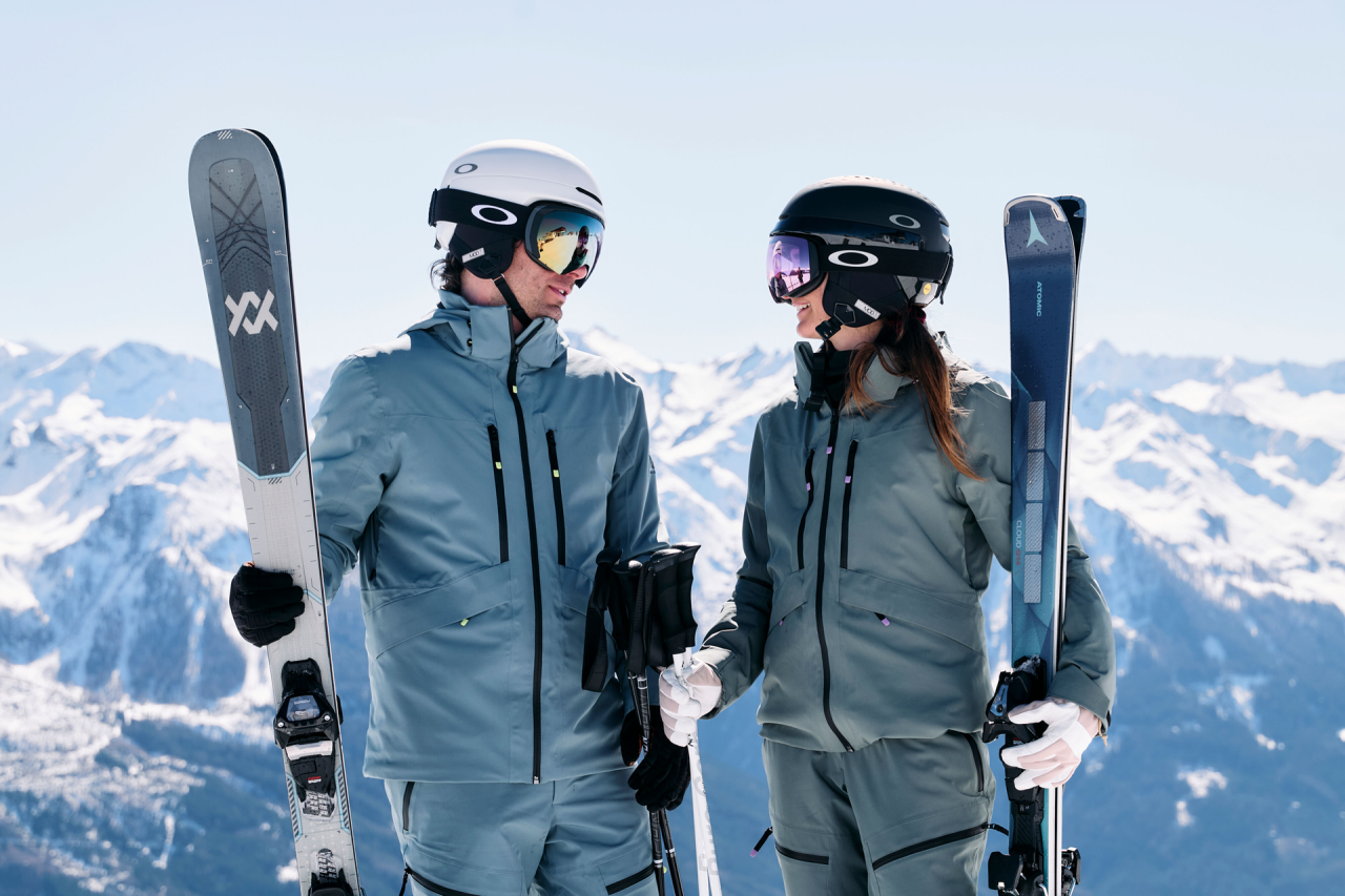 Zwei Skifahrer in passenden grauen Outfits und Helmen stehen auf einem schneebedeckten Berg, halten ihre Skier in den Händen und haben den Hintergrund von schneebedeckten Gipfeln unter einem klaren Himmel.