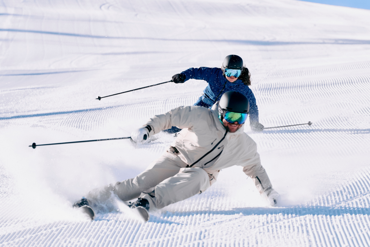 Zwei Skifahrer in Winterausrüstung gleiten einen verschneiten Hang hinunter und hinterlassen Spuren im Neuschnee. Das Sonnenlicht wirft Schatten und schafft eine dynamische, aufregende Szene.