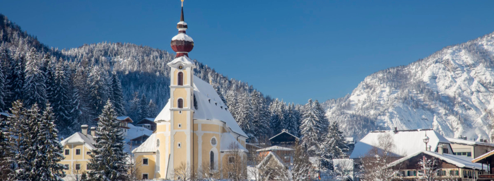 Gemütliche Dorfszene mit einer gelben Kirche, schneebedeckten Bäumen und alpinen Bergen unter einem klaren blauen Himmel, die eine ruhige Winteratmosphäre hervorruft.