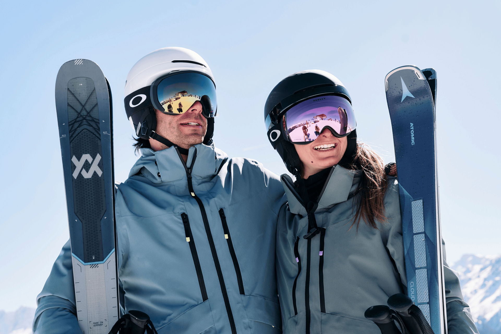 A smiling couple in ski gear holds skis against a snowy mountain backdrop. They wear helmets and goggles, conveying a sense of adventure and joy.