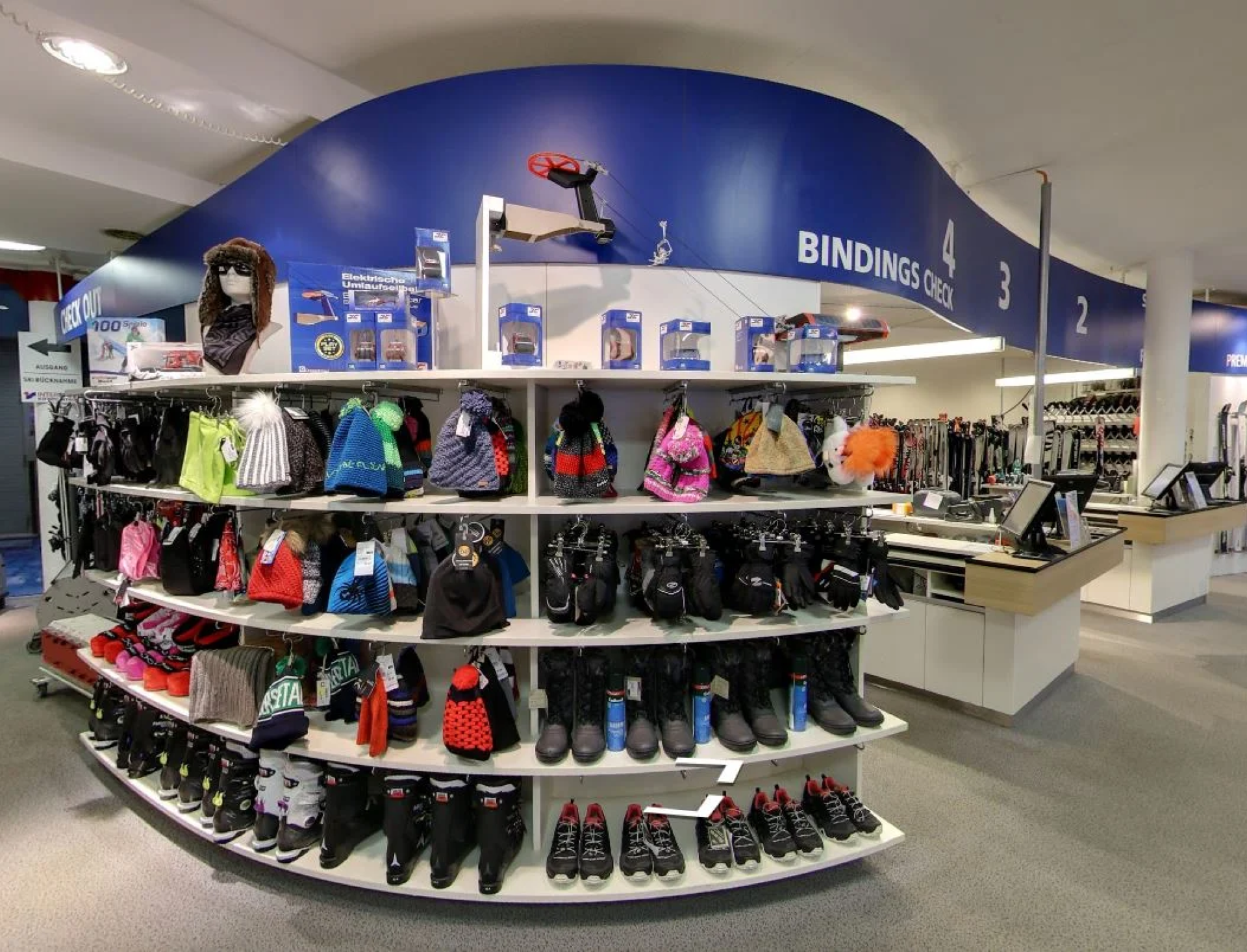 Innenraum eines Skigeschäfts mit Regalen, in denen bunte Wintermützen, Handschuhe und Skischuhe ausgestellt sind. Blaue Beschilderungen heben verschiedene Bereiche hervor, um die Orientierung zu erleichtern.