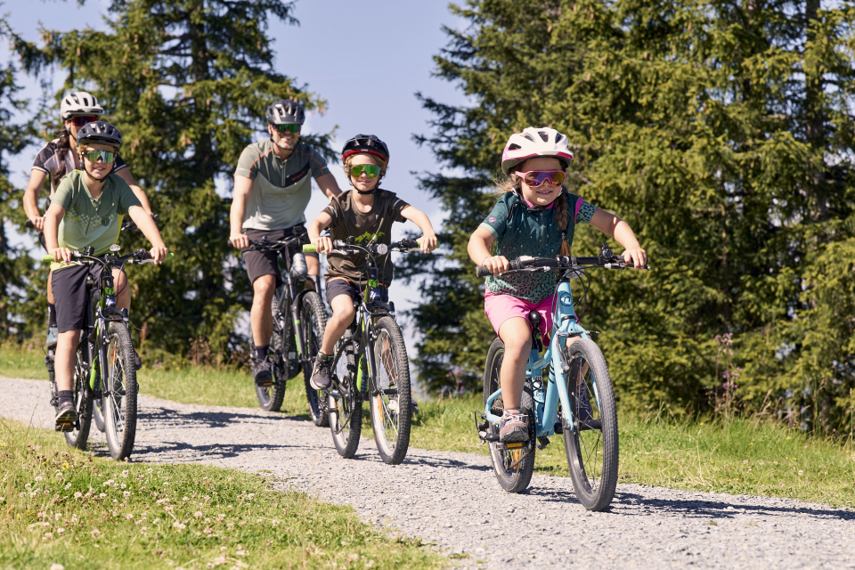 A group of five cyclists, including children and adults, ride on a gravel path through a lush green forest, all wearing helmets and sunglasses, smiling.