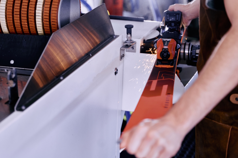 Ein Skiservice-Mitarbeiter schleift einen orangefarbenen Ski an einer Maschine, Funken sprühen vom Metall.