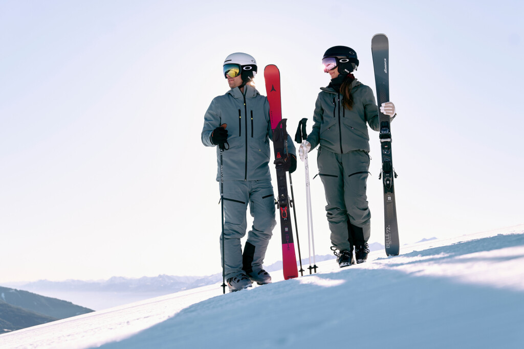 Zwei Skifahrer in grauer Ausrüstung stehen mit Skiern in der Hand auf einer sonnigen Piste und lächeln sich an.