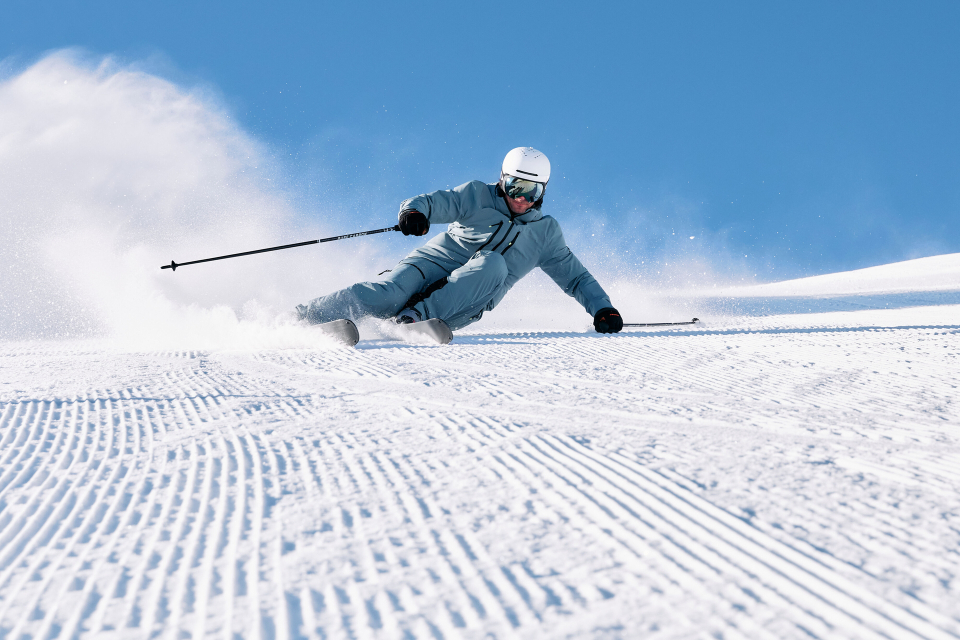 Skifahrer in blauer Kleidung und weißem Helm, der gekonnt eine schneebedeckte Piste unter strahlend blauem Himmel hinunterfährt, während hinter ihm eine Schneewolke aufwirbelt.
