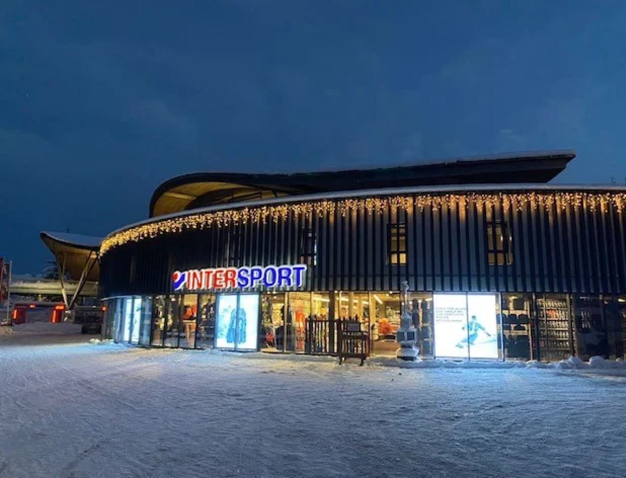 Außenansicht eines modernen Gebäudes in der Dämmerung, mit einem hell erleuchteten „INTERSPORT“-Schild und Eiszapfenlichtern. Schnee bedeckt den Boden und schafft eine ruhige Winterlandschaft.