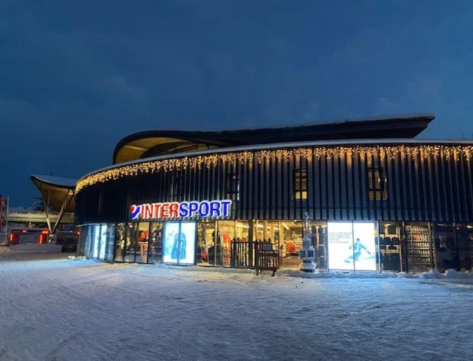 Außenansicht eines modernen Gebäudes in der Dämmerung, mit einem hell erleuchteten „INTERSPORT“-Schild und Eiszapfenlichtern. Schnee bedeckt den Boden und schafft eine ruhige Winterlandschaft.