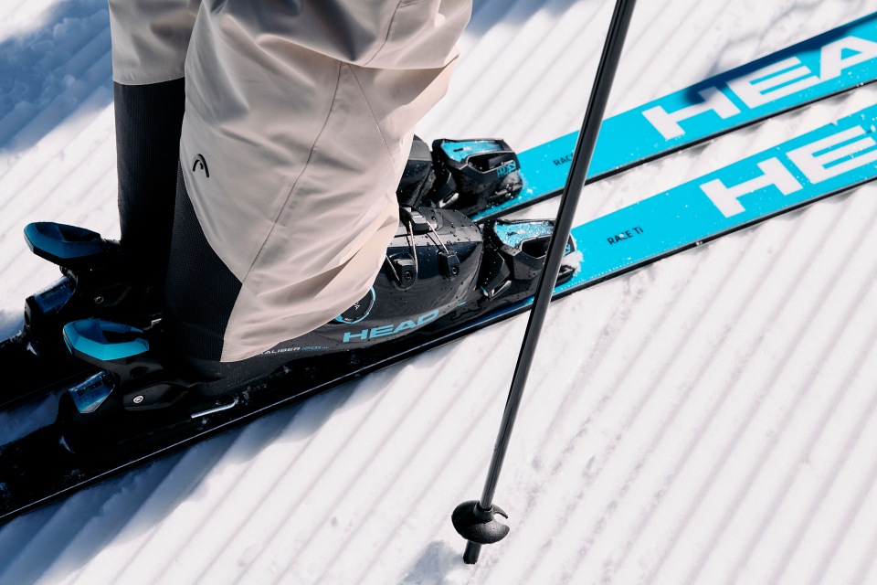 A skier wearing beige pants and black boots stands on blue and black skis on groomed snow, holding a ski pole in his hand. The scene conveys a sense of adventure.