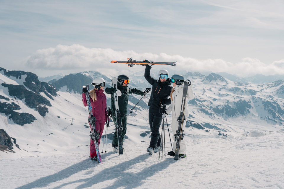 Drei Skifahrer in farbenfroher Ausrüstung stehen auf einem schneebedeckten Berggipfel, einer von ihnen reckt triumphierend seine Skier in die Höhe. Majestätische, schneebedeckte Berge umgeben sie.