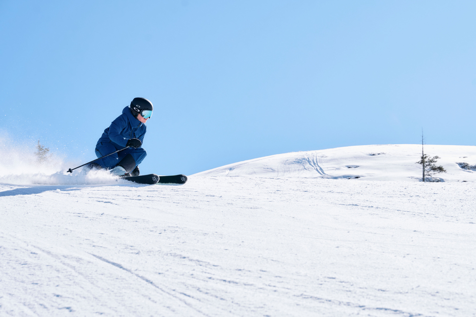 Ein Skifahrer in blauer Ausrüstung fährt unter strahlend blauem Himmel rasant einen verschneiten Hang hinunter. Die Szene vermittelt die Aufregung und den Nervenkitzel des Wintersports.